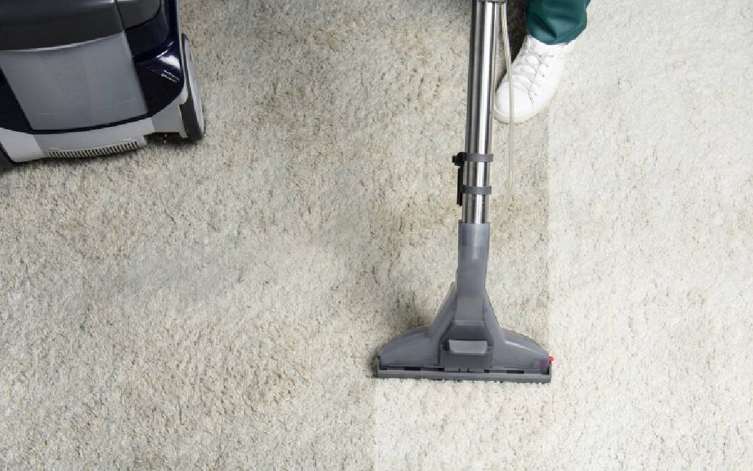 A top-down overhead shot showing a striking before-and-after contrast on a high-pile cream carpet during a professional cleaning. A vacuum extractor wand is pulling up dirt, leaving a stark, bright white cleaned path next to the uncleaned, duller section of the rug.