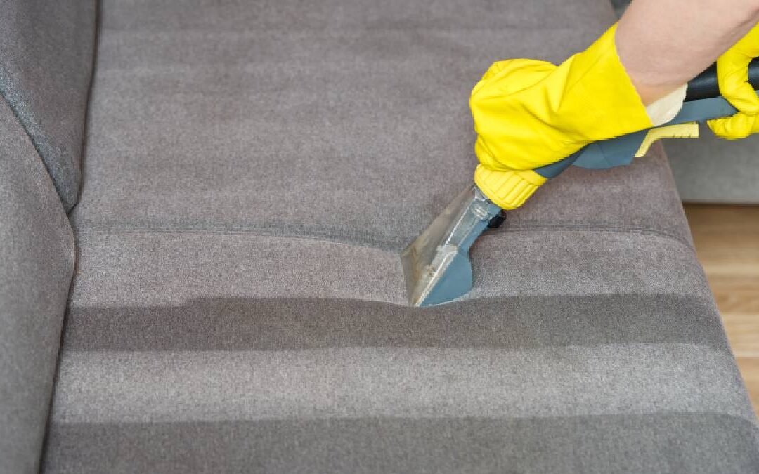 A close-up shot of a person wearing bright yellow rubber gloves using a handheld upholstery extraction wand to clean a grey fabric sofa. The clear nozzle leaves distinct, parallel cleaning lines across the cushion's textured surface.